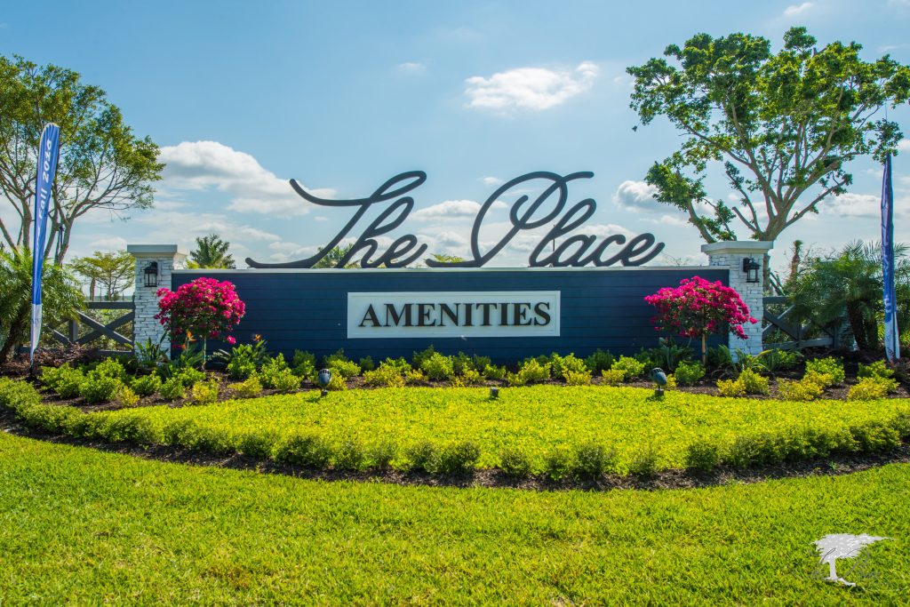The Place amenities entrance sign with landscaped flowers and trees under a blue sky.