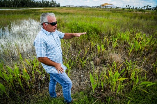 A man wearing sunglasses points toward a grassy marshland landscape under an overcast sky.