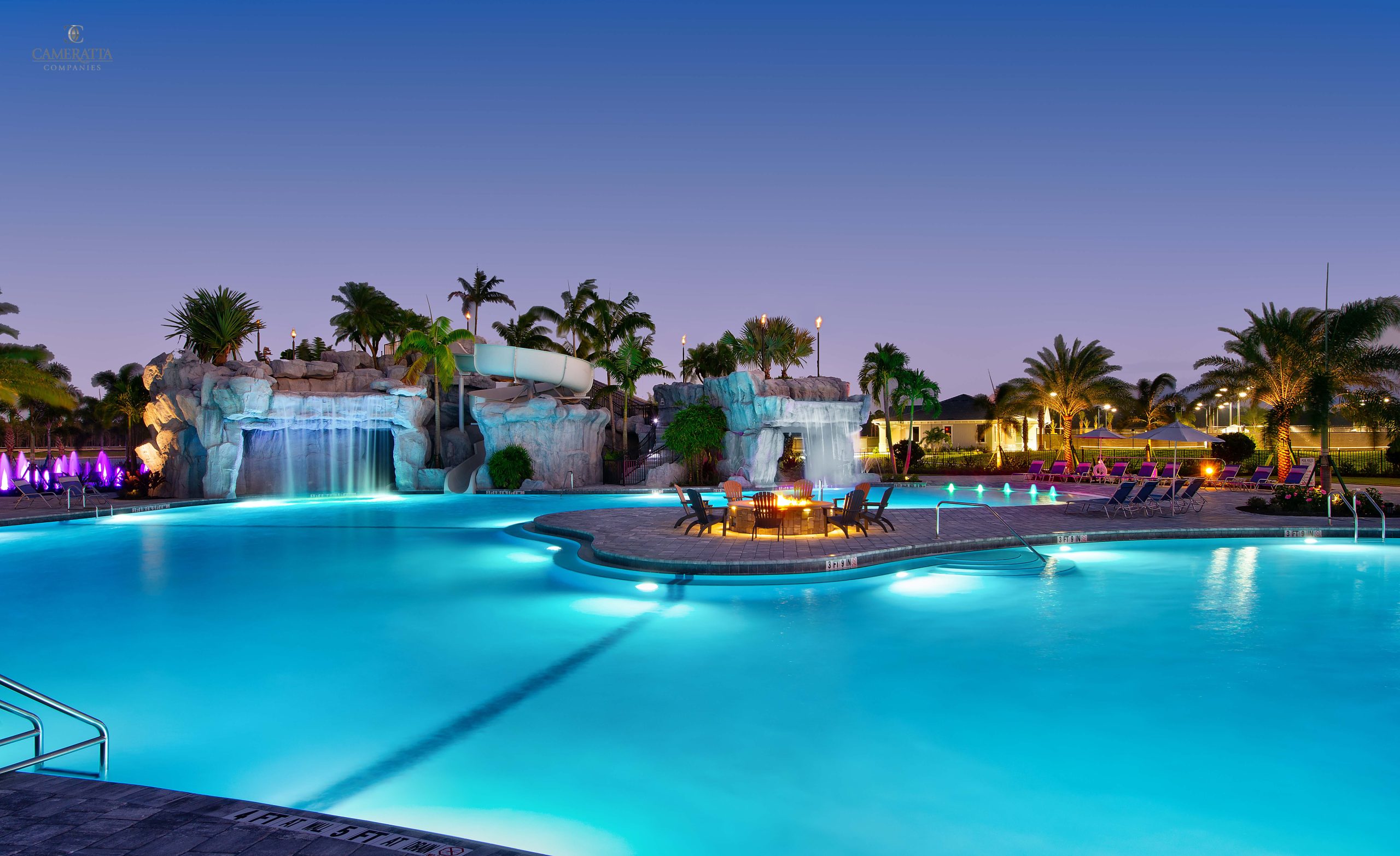 Swimming pool resort at dusk with palm trees and colorful lights reflecting on the water.