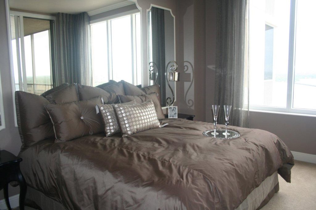 Spacious bedroom with a large bed, gray bedding, and pillows in front of tall windows with curtains.