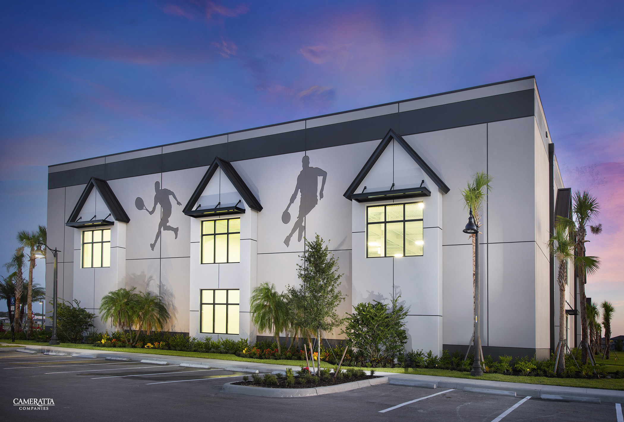 Modern white building with triangular window frames and hanging silhouettes against a purple sunset sky.