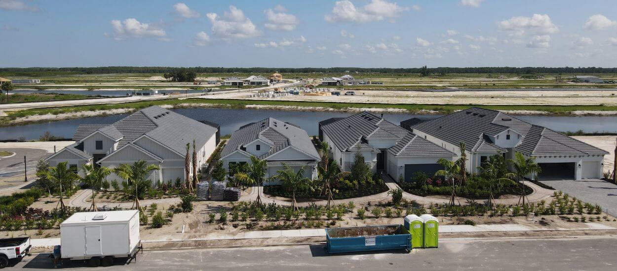 Aerial view of a row of gray-roofed homes beside a road and construction bins, with wetlands in the distance.